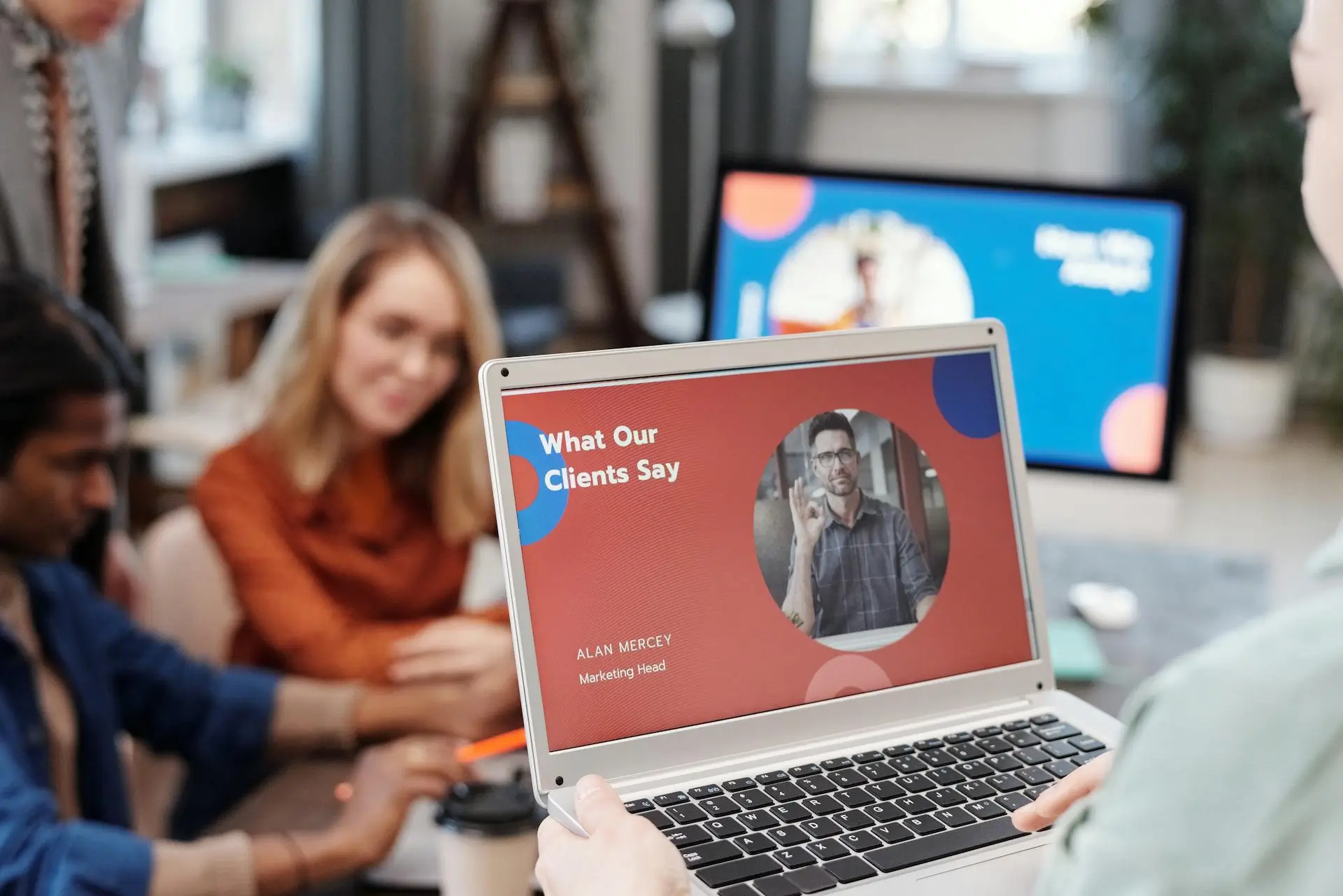 A person holds a laptop showing a presentation slide titled What Our Clients Say with a photo of a man and the name Alan Mercey, Marketing Head. In the background, three people are seated at a table, having a discussion.