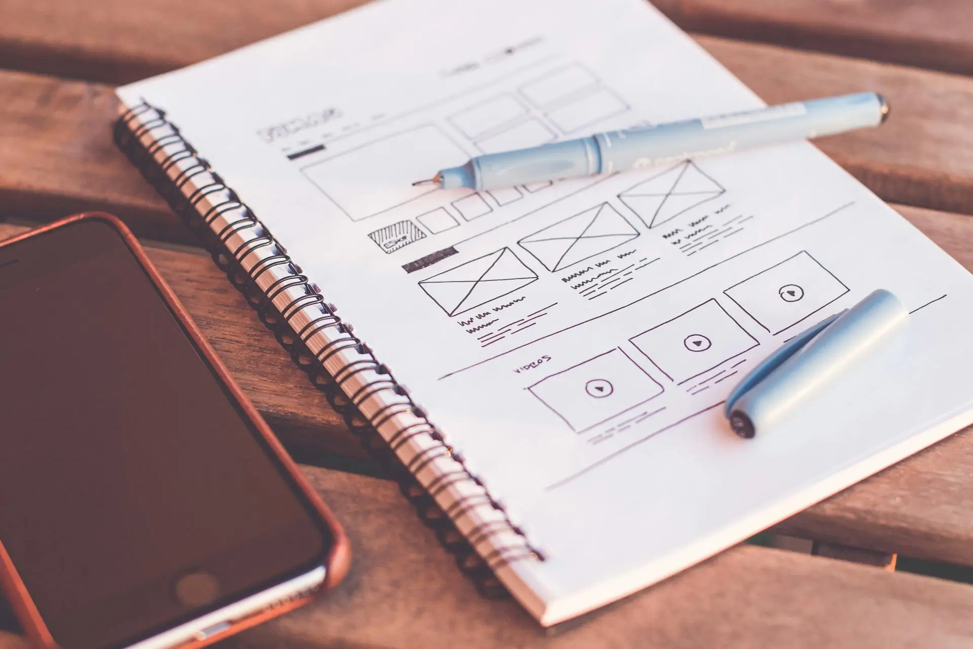 A spiral notebook displaying website wireframes sits on a wooden table. A blue pen rests on the open notebook, and a smartphone lies nearby. The tables natural texture adds a warm, rustic feel to the scene.