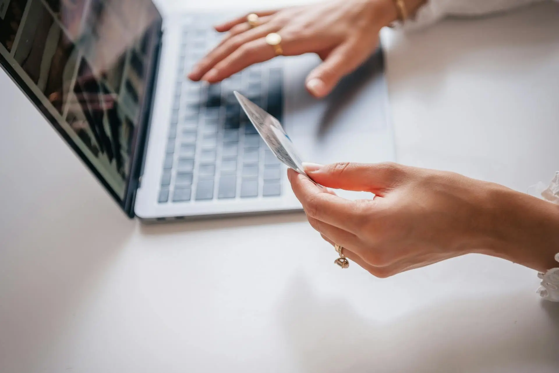 A person holding a credit card in one hand while using a laptop with the other, suggesting online shopping or payment. The hands are adorned with rings. The laptop is on a white surface.