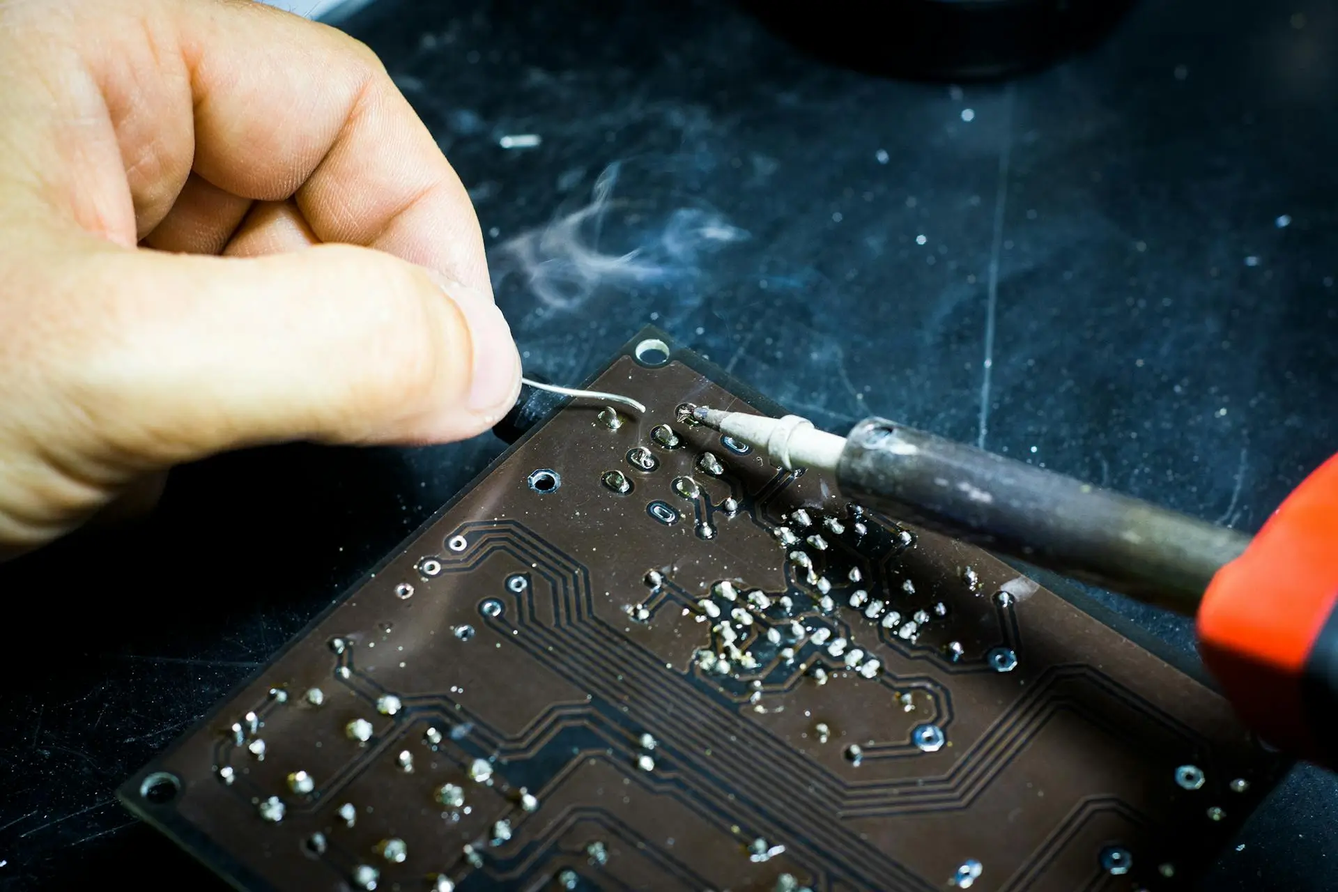 Close-up of a hand soldering a circuit board on a dark surface. Smoke rises from the heated area as a soldering iron and solder wire are used to connect components, highlighting the intricate electronic work.