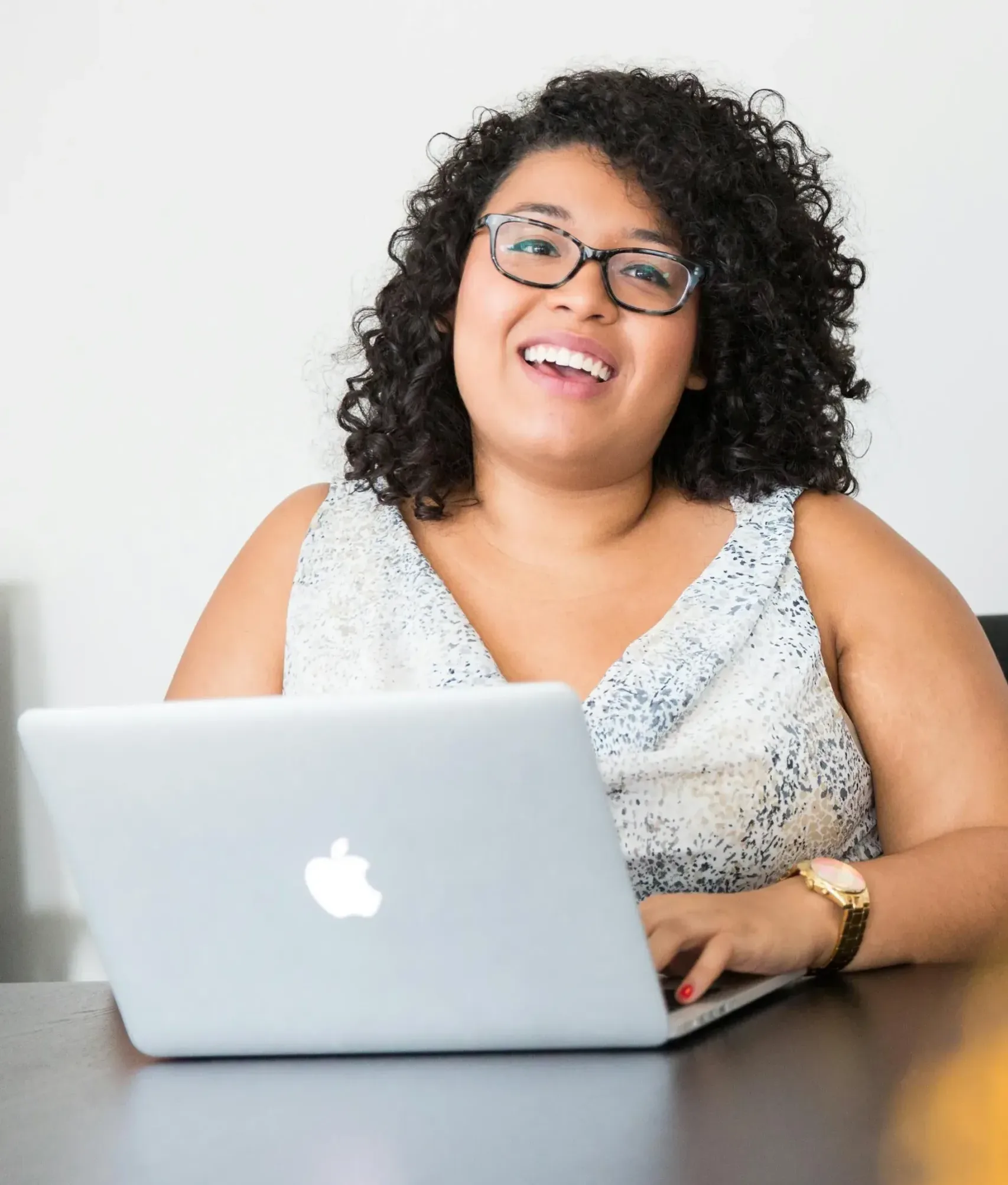 A women with curly hair and glasses is smiling while sitting at a desk using a laptop. The laptop is silver with an apple logo. They are wearing a patterned sleeveless top.