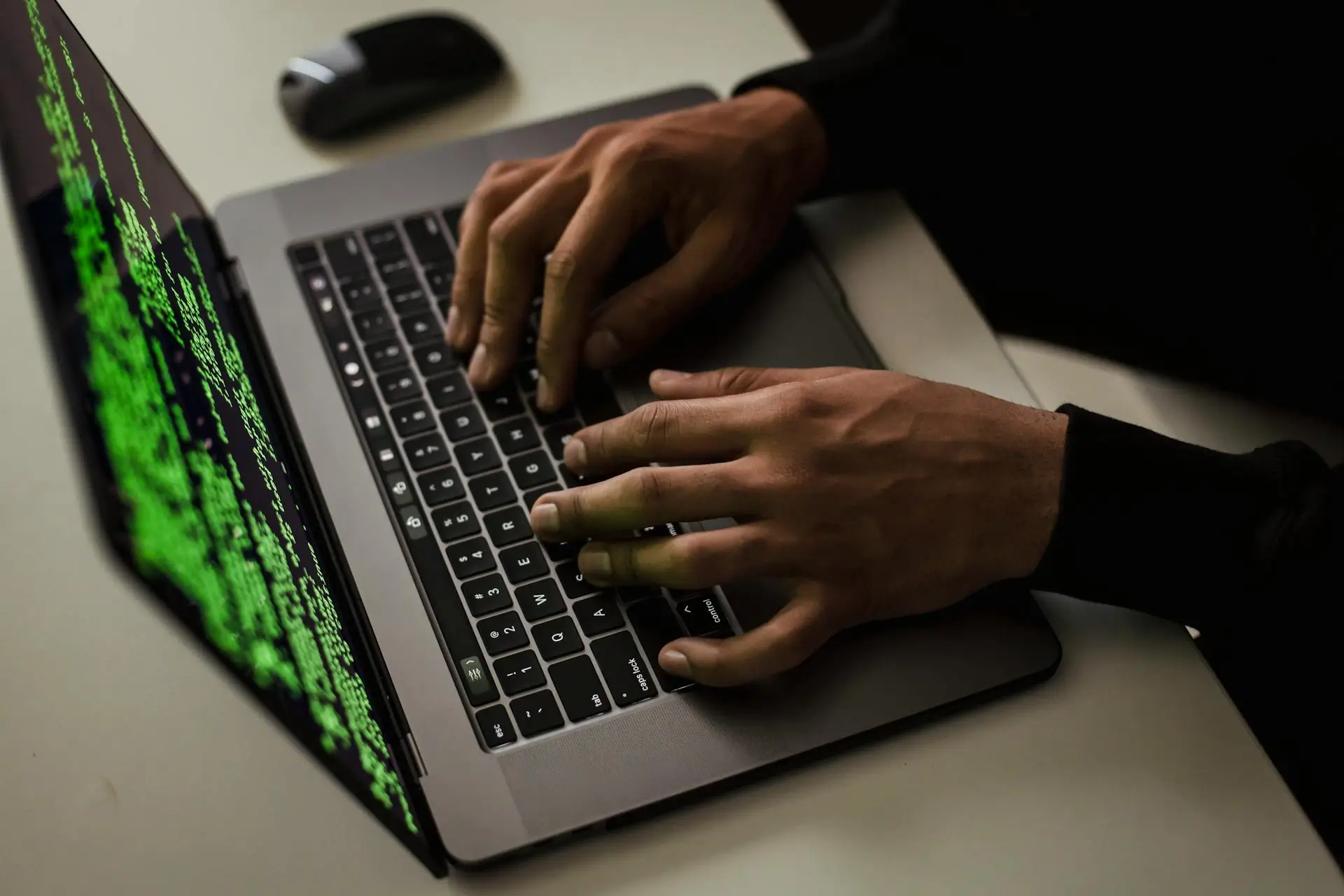 Person typing on a laptop keyboard with green code text displayed on the screen.