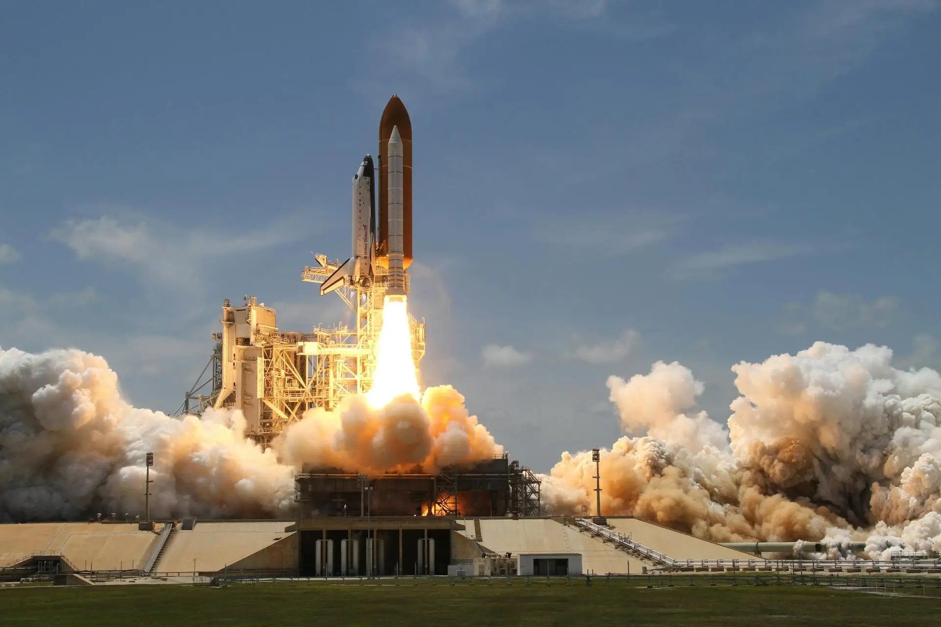 A space shuttle is launching from a pad, with bright flames and smoke billowing out from its engines. The sky is clear and blue, and the launch pad structure is visible, surrounded by a grassy field.