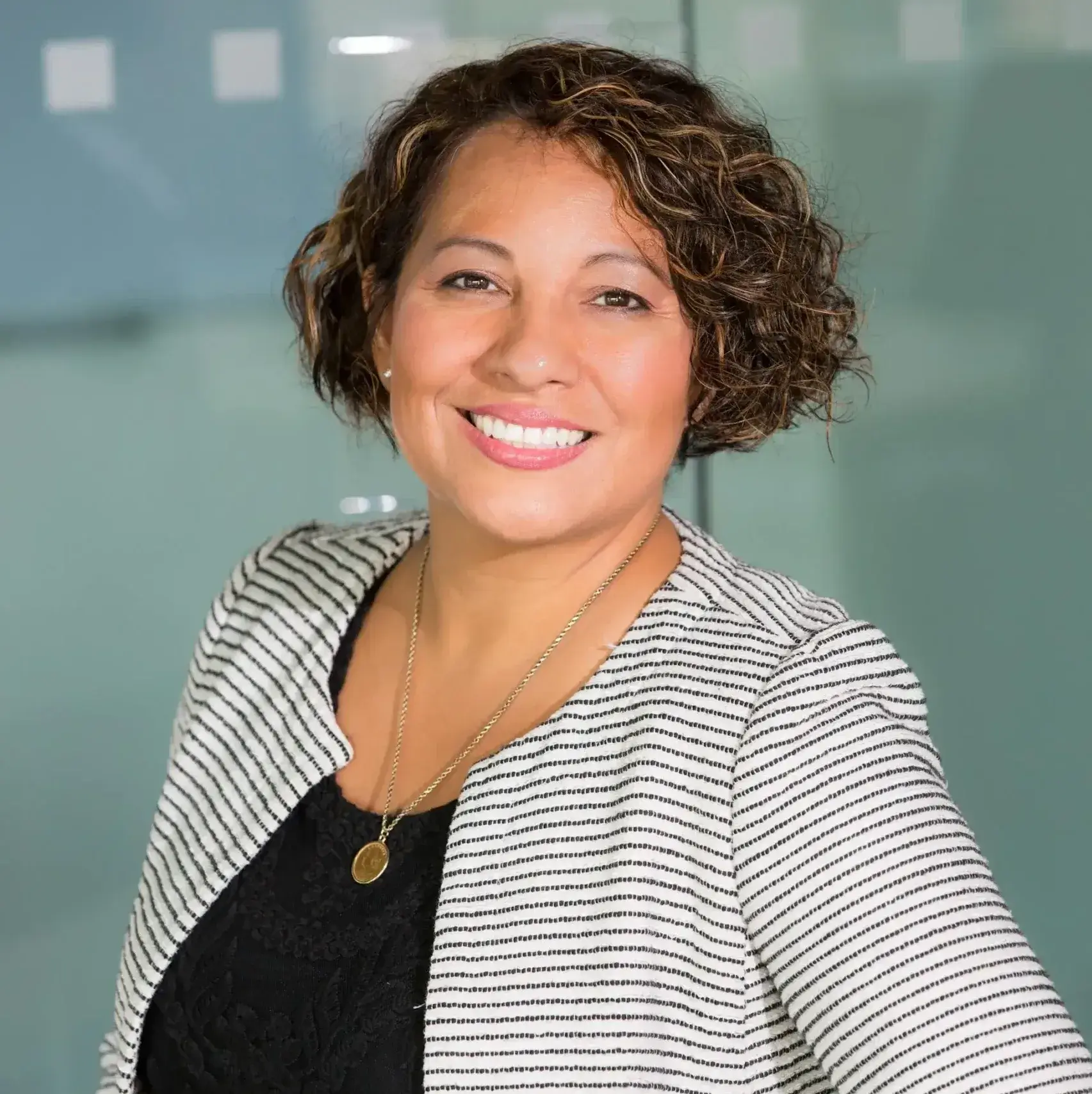 A women with curly hair smiles while wearing a striped blazer and dark shirt, standing against a blurred glass background.