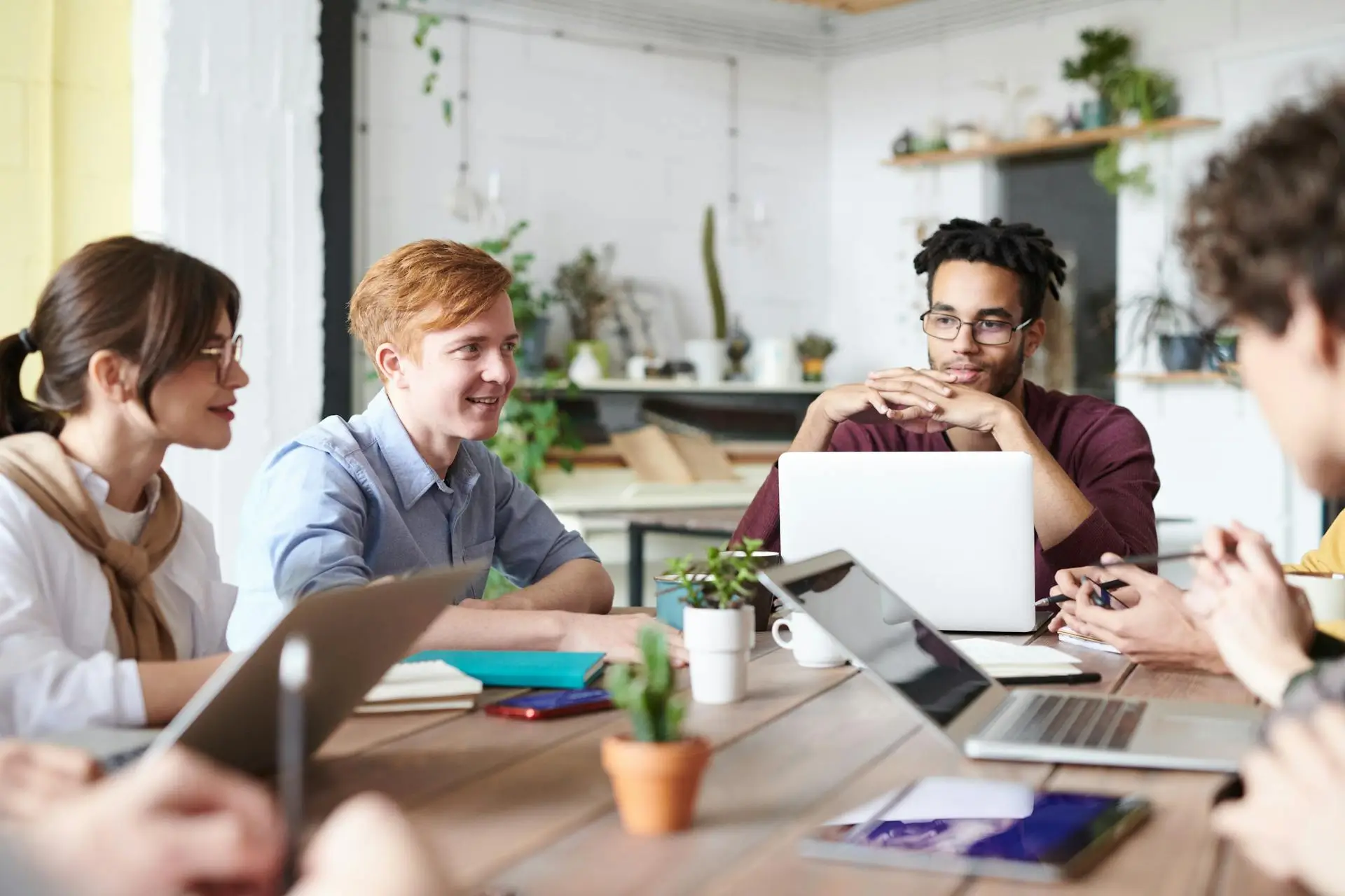 A diverse group of people engaged in a meeting around a large table with laptops and notebooks. The room has a casual atmosphere with plants and shelves in the background. Everyone appears focused and attentive.