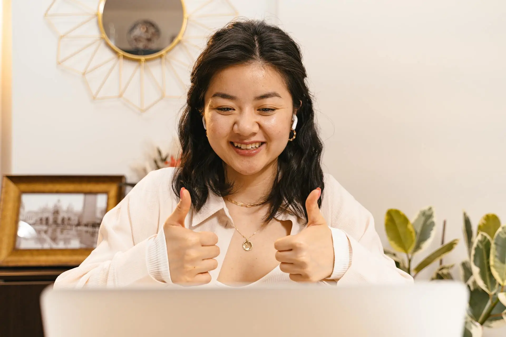 A woman with earbuds and a warm smile gives a thumbs-up gesture while sitting at a table. Behind her is a wall with a decorative mirror and a framed photo, alongside some green plants.