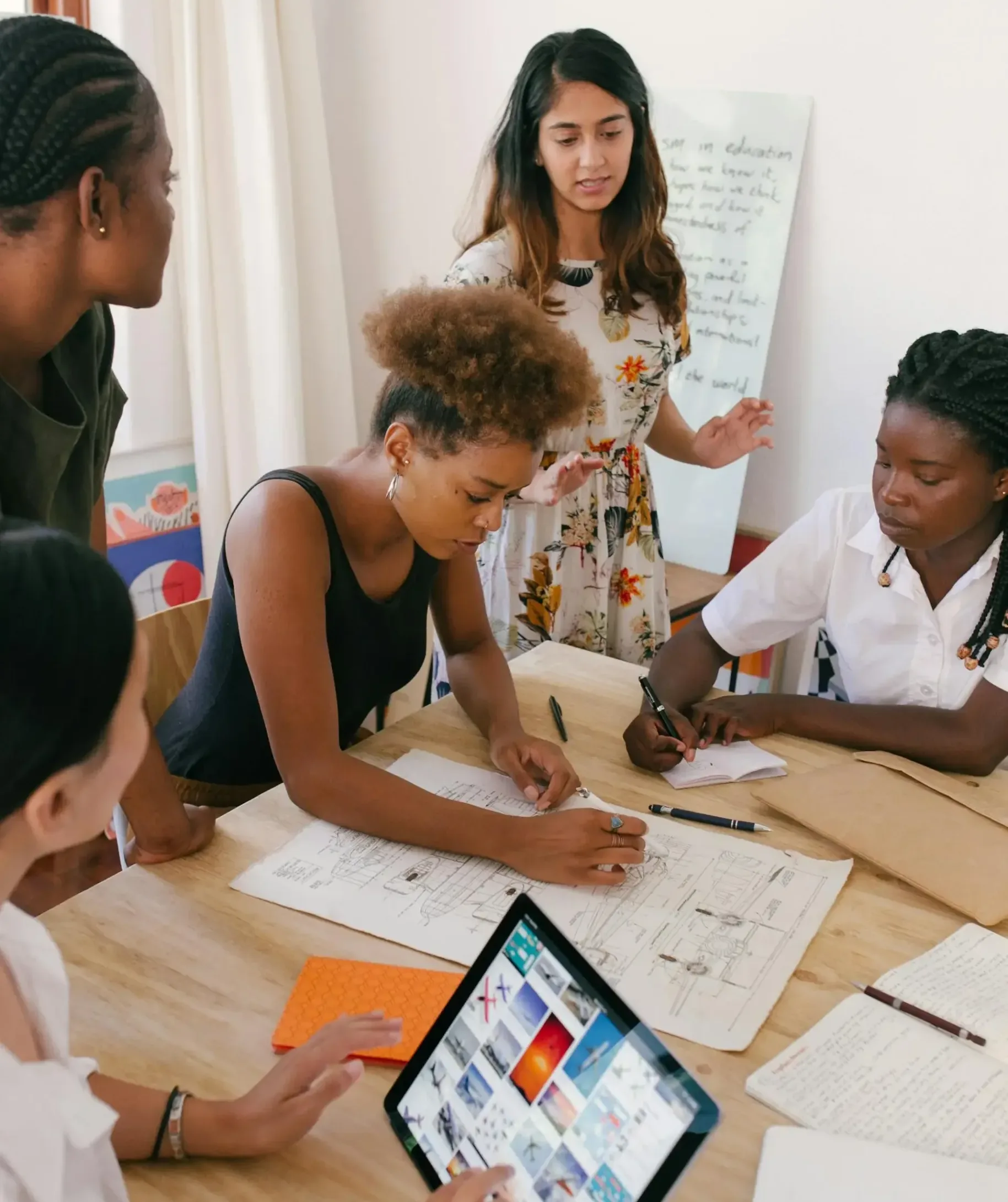 Five people collaborating around a table covered with papers and a tablet. One person is standing, gesturing towards the group. Others are seated, actively discussing and taking notes. A whiteboard with notes is visible in the background.