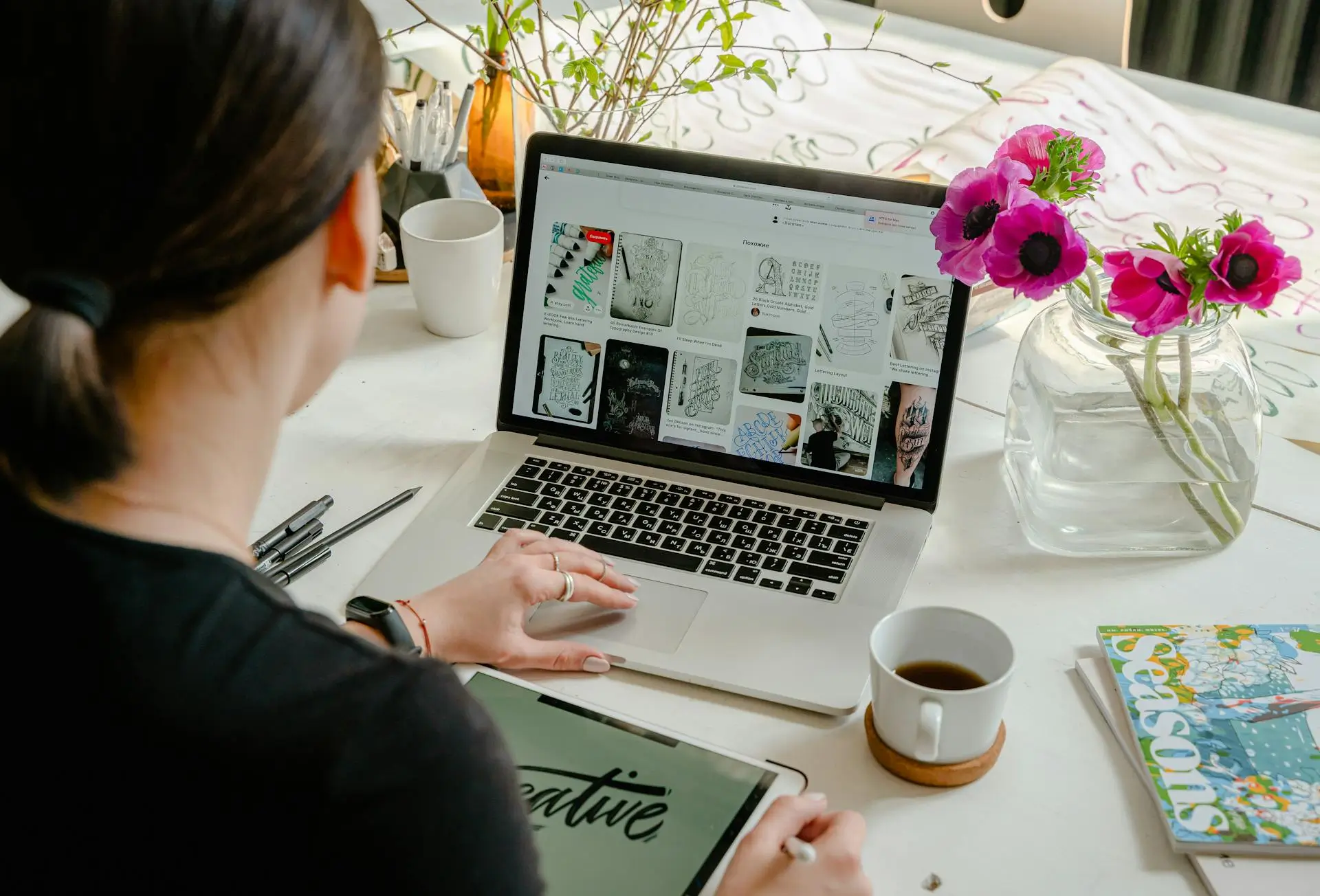 A person sits at a table, using a laptop that displays various sketch images. Nearby are a tablet with the word creative, a cup of coffee, a magazine, and a vase with purple flowers. Pens and a cup are also on the table.