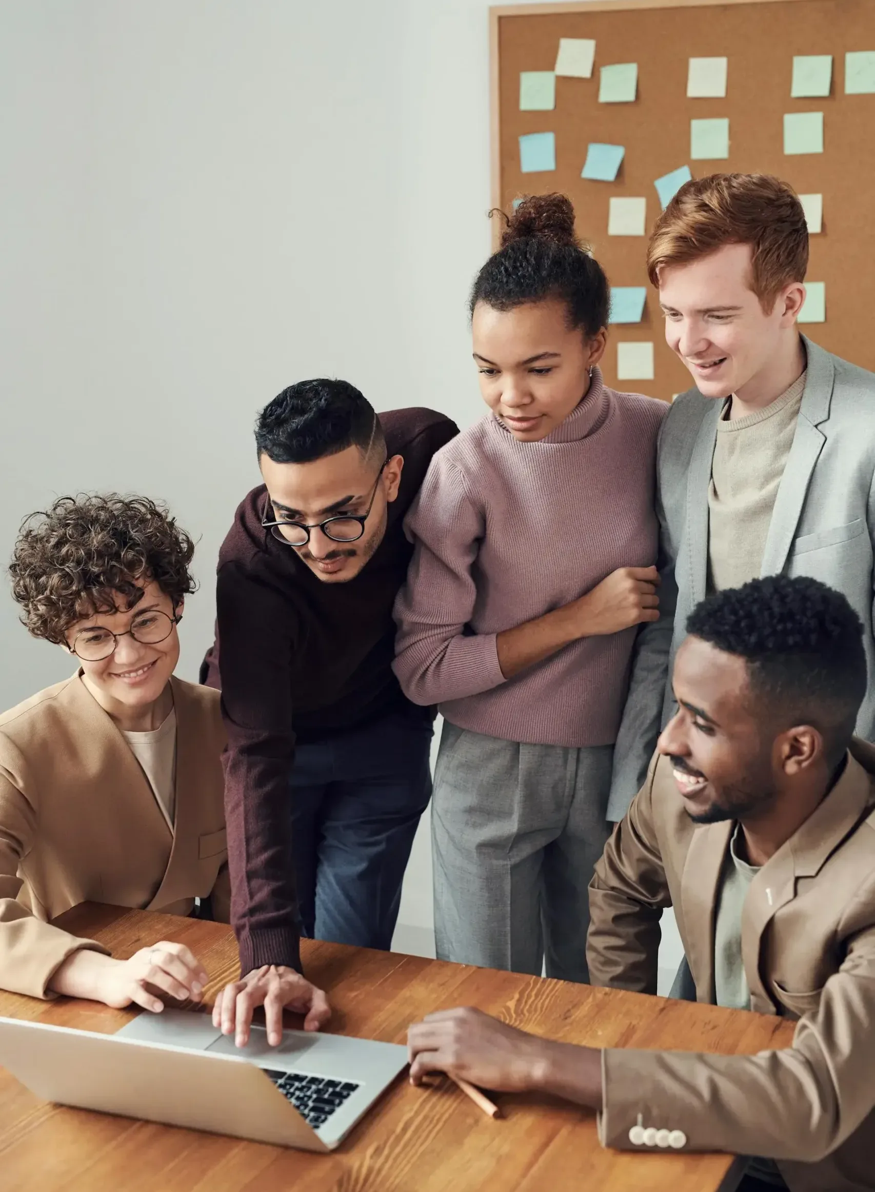 A diverse group of five people gathers around a laptop, engaged in discussion. They are in an office setting with a corkboard and sticky notes in the background. The group appears collaborative and focused.